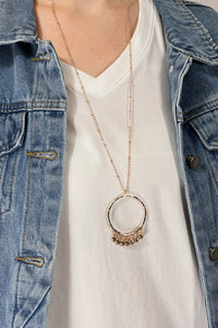 Model is wearing a long gold chain necklace with a hoop pendant that has fringe. 