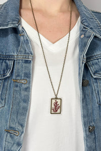 Model is wearing a long brass chain necklace with a pressed flower pendant. 