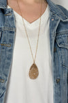 Model is wearing a long gold chain necklace with a teardrop shaped pendant that features rows of shiny small beads. 