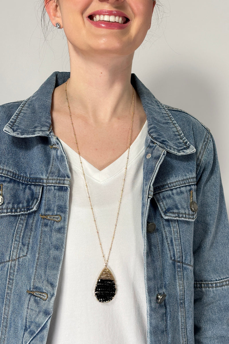 Model is wearing a long gold chain necklace with a teardrop shaped pendant that features rows of shiny small beads. 