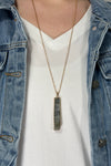 Model is wearing a long gold chain necklace with a vertical rectangle pendant in a grey color. 