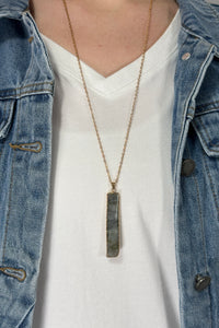 Model is wearing a long gold chain necklace with a vertical rectangle pendant in a grey color. 