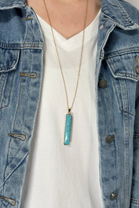 Model is wearing a long gold chain necklace with a vertical rectangle pendant in a turquoise color. 