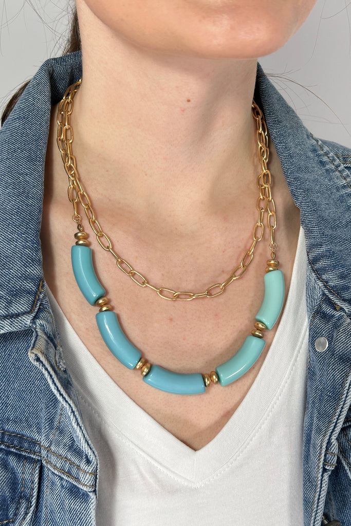 Model is wearing a double layer necklace. The first layer features a brushed gold chain link and the second layer has chunky teal colored beads. 