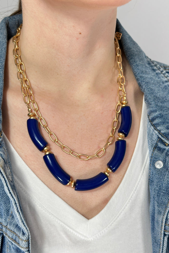 Model is wearing a double layer necklace. The first layer features a brushed gold chain link and the second layer has chunky royal blue colored beads. 