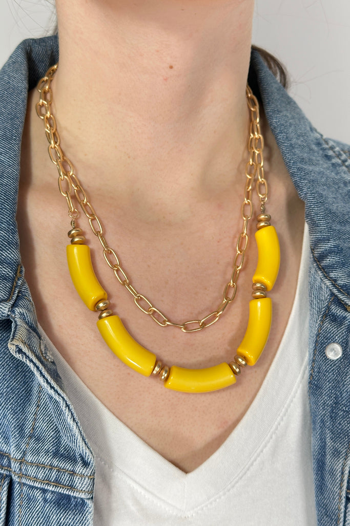 Model is wearing a double layer necklace. The first layer is a brushed gold chain link and the second features large yellow beads. 