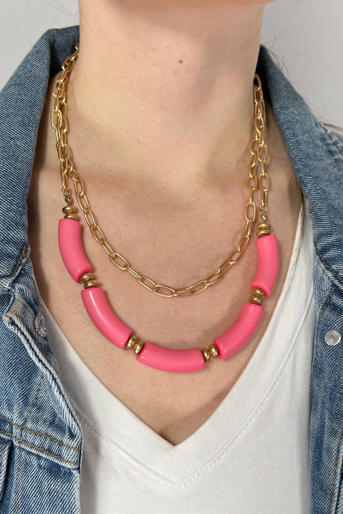 Model is wearing a double layer necklace. The first layer features a brushed gold chain link and the second layer has chunky pink colored beads. 