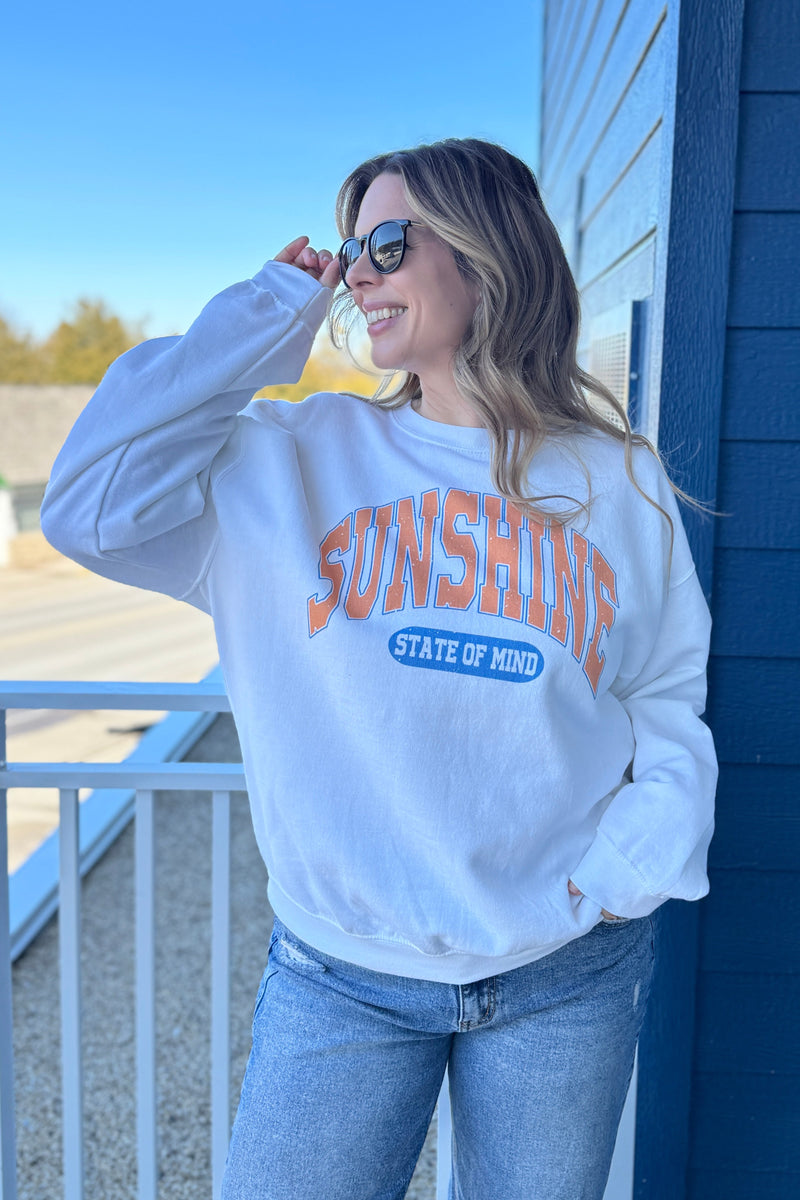 Woman wearing a white sweatshirt with 'Sunshine State of Mind' text, standing on a balcony.