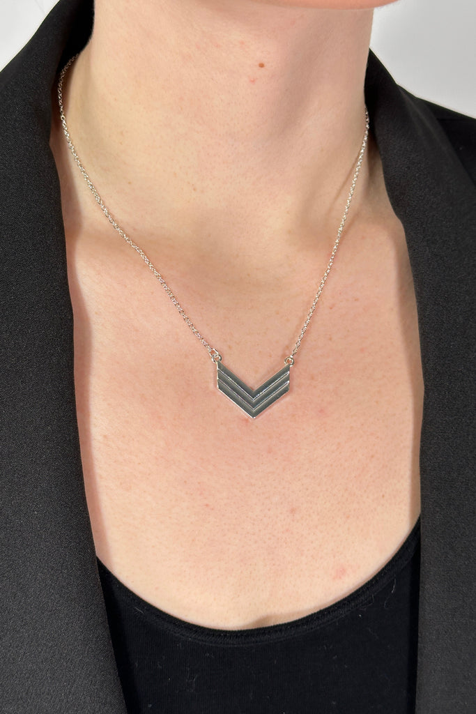 Model is wearing a collar length silver chain necklace that features a small arrow pendant. 