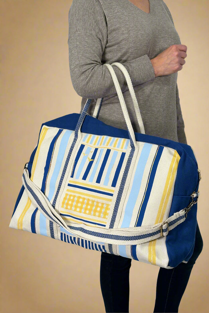 Model is holding a large canvas weekender bag with nautical stripes. 