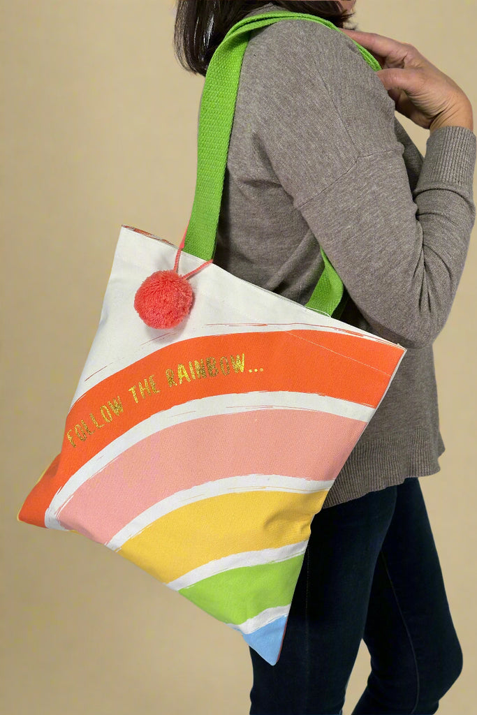 Model is holding a rainbow canvas tote bag with a pom on the strap.