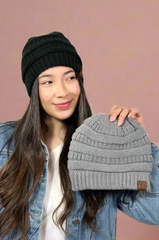 Model is wearing a black cable knit beanie that is lined and holding up the same beanie available in grey. 