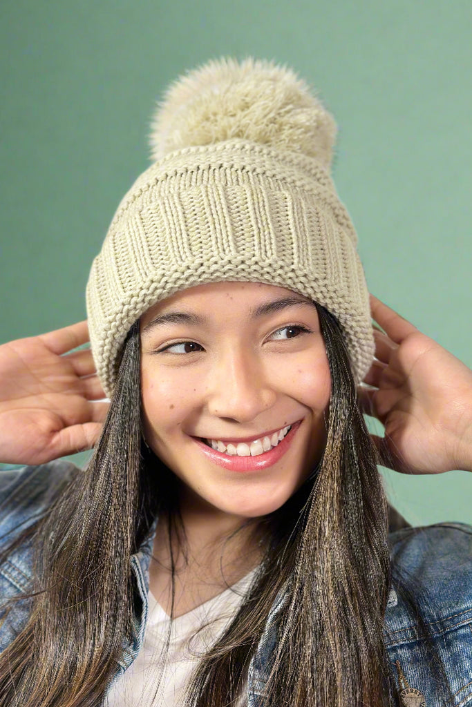 Model is wearing a cream colored slouchy cable knit beanie with a large pom pom on top.