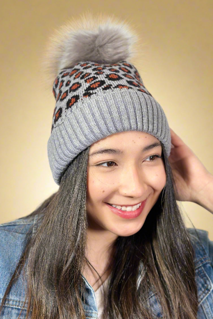 Model is wearing a grey leopard printed hat that features a grey faux fur pom on top.