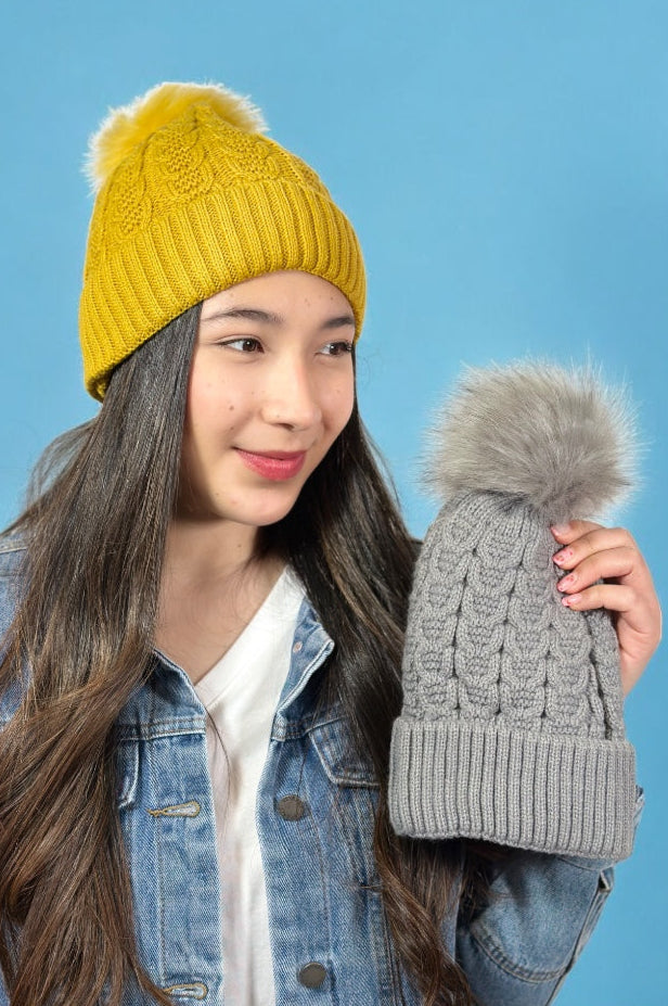 Model is wearing a mustard colored hat with a faux fur pom on top. She's holding the same hat available in grey.