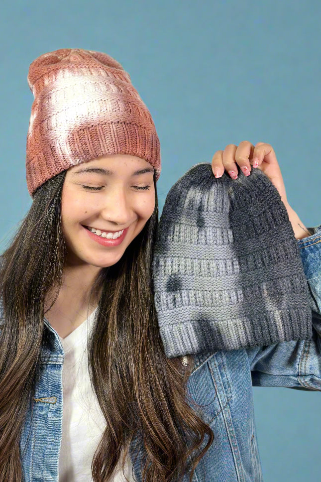 Model is wearing a peachy colored tie dye knit beanie and holding up the same beanie in a grey color way.