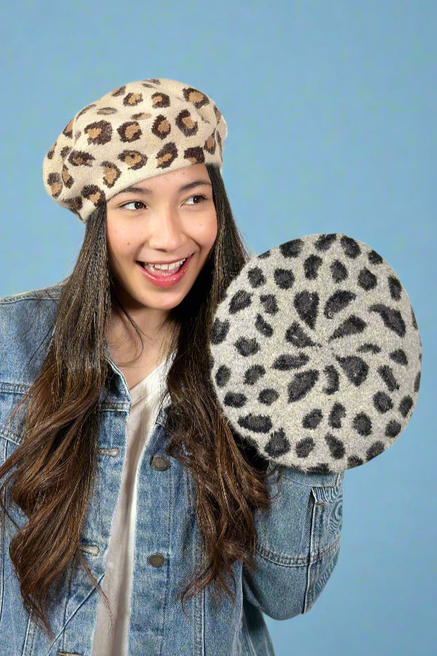 Model is wearing a leopard print beret in a cream color way and holding up the same beret but in a grey color way. 