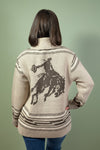 Person wearing a beige cardigan with a graphic of a person on a horse, standing against a plain background.