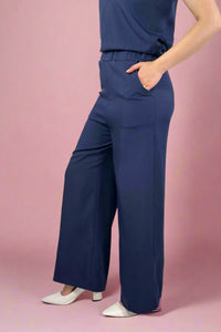Woman wearing a black sleeveless top and gray pants standing indoors with a window and plant in the background.