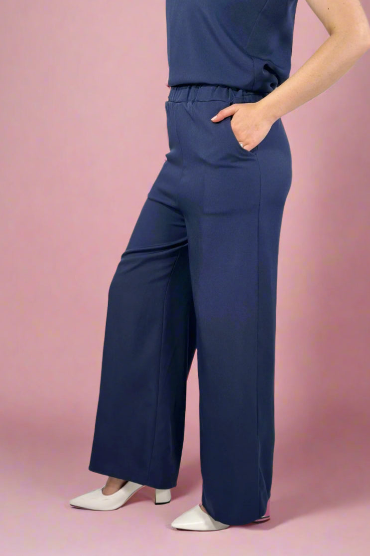 Woman wearing a black sleeveless top and gray pants standing indoors with a window and plant in the background.