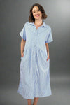 Woman wearing an oversized striped cream and light blue button down midi dress with pockets. Cuffed short sleeves.