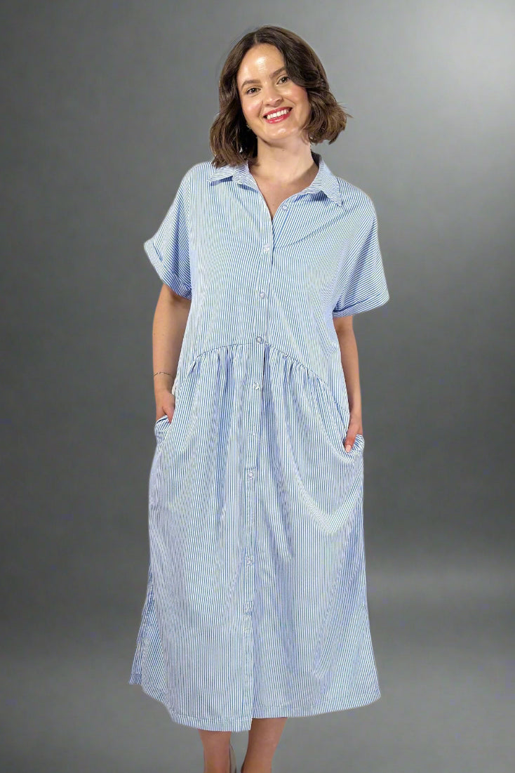 Woman wearing an oversized striped cream and light blue button down midi dress with pockets. Cuffed short sleeves.