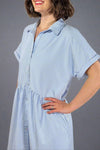 Woman wearing an oversized striped cream and light blue button down midi dress with pockets. Cuffed short sleeves.