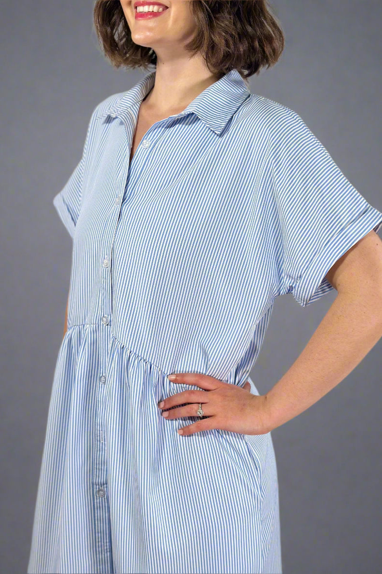 Woman wearing an oversized striped cream and light blue button down midi dress with pockets. Cuffed short sleeves.