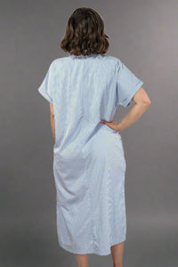 Woman wearing an oversized striped cream and light blue button down midi dress with pockets. Cuffed short sleeves.