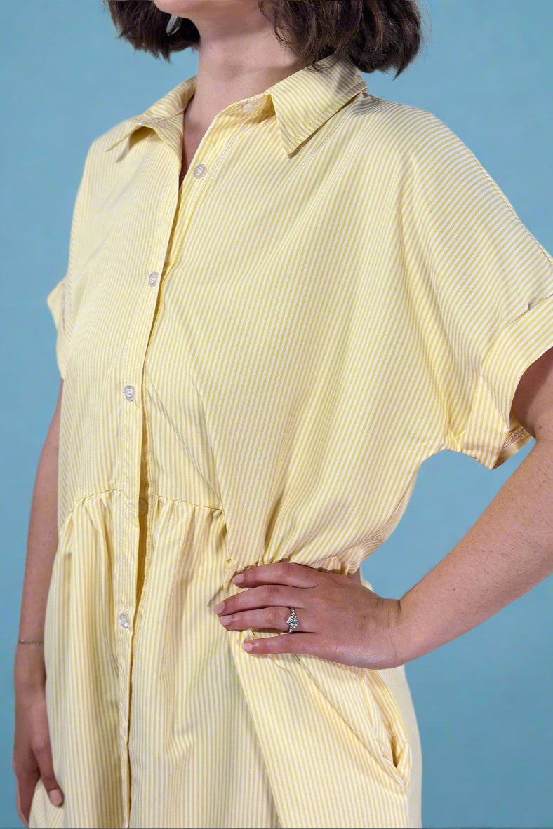 Woman wearing an oversized striped cream and yellow button down midi dress with pockets. Cuffed short sleeves.