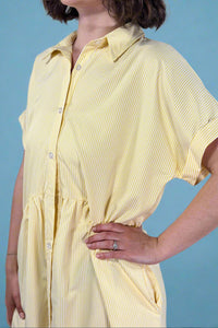 Woman wearing an oversized striped cream and yellow button down midi dress with pockets. Cuffed short sleeves.