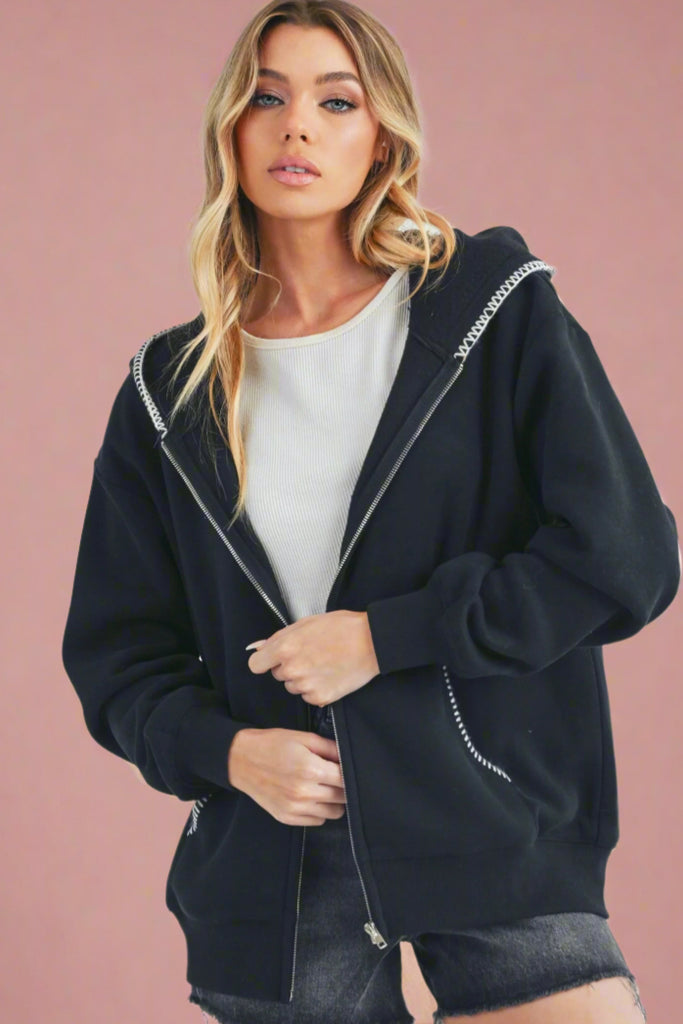 Model is wearing a classic black zip up hoodie with an oversized fit. Contrast white stitching on the hood and pockets add a playful and trendy touch. 