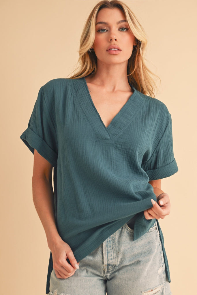 Model is wear a loose casual v neck top in teal. The top features rolled cuff sleeves, a raw edge v-neck. Split hem that is longer in the back. 