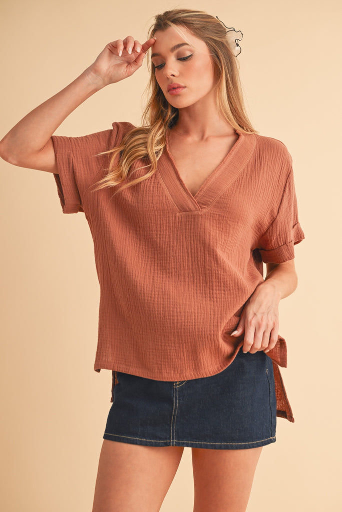 Model is wearing a casual short sleeve top in a terracotta color. The top features cuffed short sleeves, a raw v-neck and a longer hemline in back. 