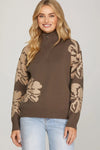 Woman wearing a brown sweater with beige floral patterns on sleeves against a plain background