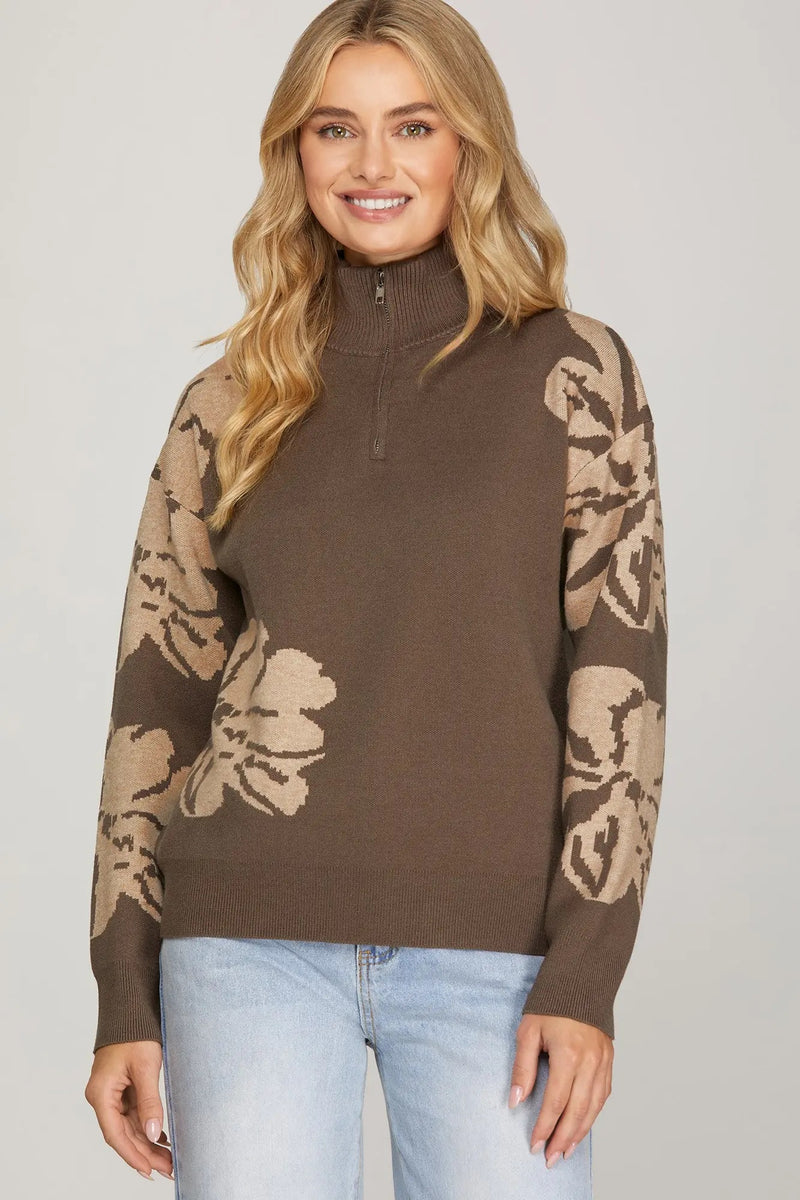 Woman wearing a brown sweater with beige floral patterns on sleeves against a plain background