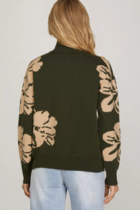 Person wearing a dark green sweater with beige floral patterns on sleeves against a plain background
