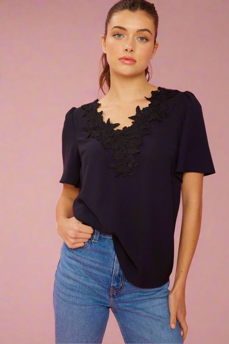 A women’s solid short sleeve blouse In black featuring lace overlay v-neckline.