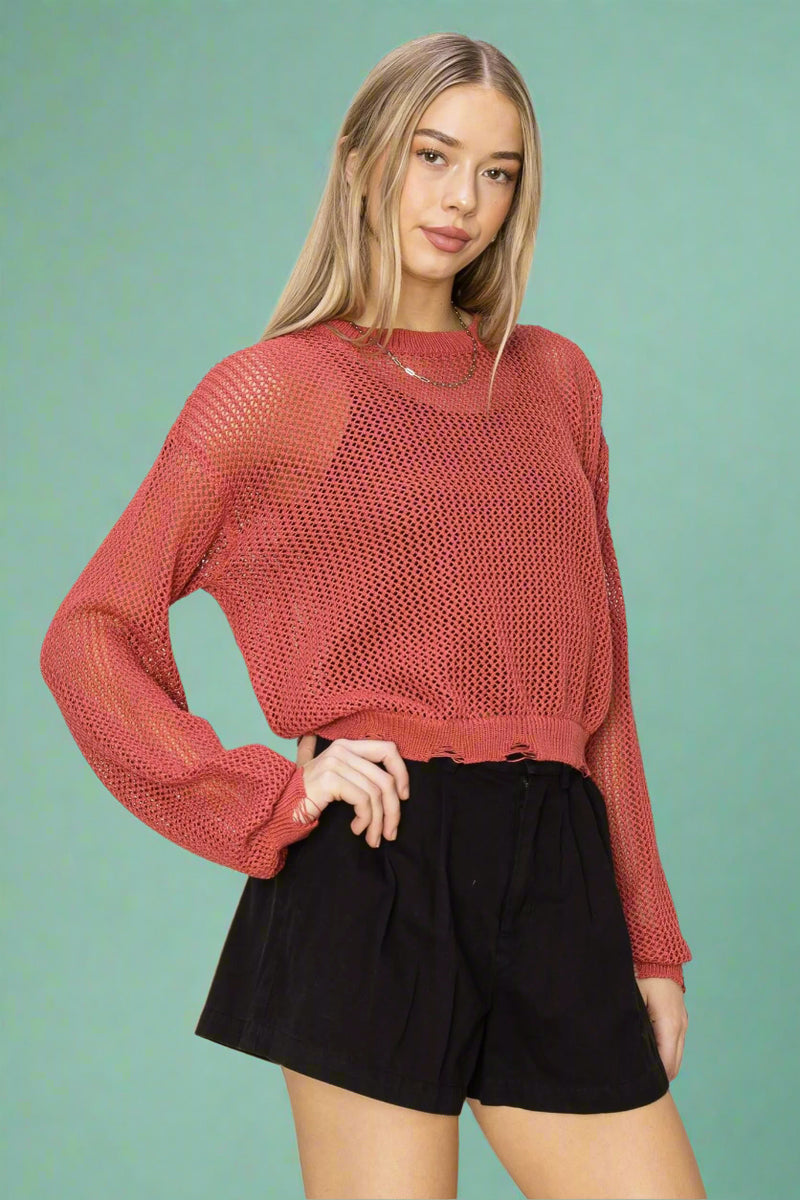 Model is wearing a loose knit cropped rust colored sweater featuring distressed detailing on the cuffs and along with hem. 