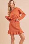 This mini skirt is an ochre orange. It has a stretch waistband, side pockets, and a ruffle hem. It is made from soft fabric.