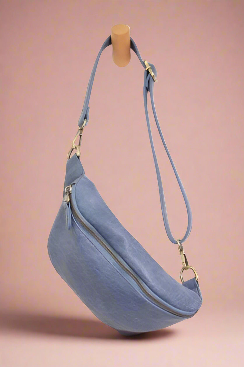 A sling bag in the color denim. It has a zipper enclosure and brass accents.
