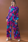 A women’s long sleeve wide leg jumpsuit featuring banded waist, zipper back, pleated wide leg fit, and vibrant leaf print in fuchsia, blue, teal, and purple.