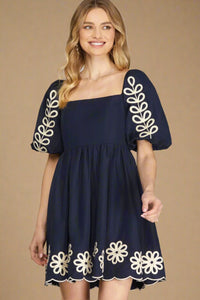 Navy dress with white floral embroidery on sleeves and hem against a tan background