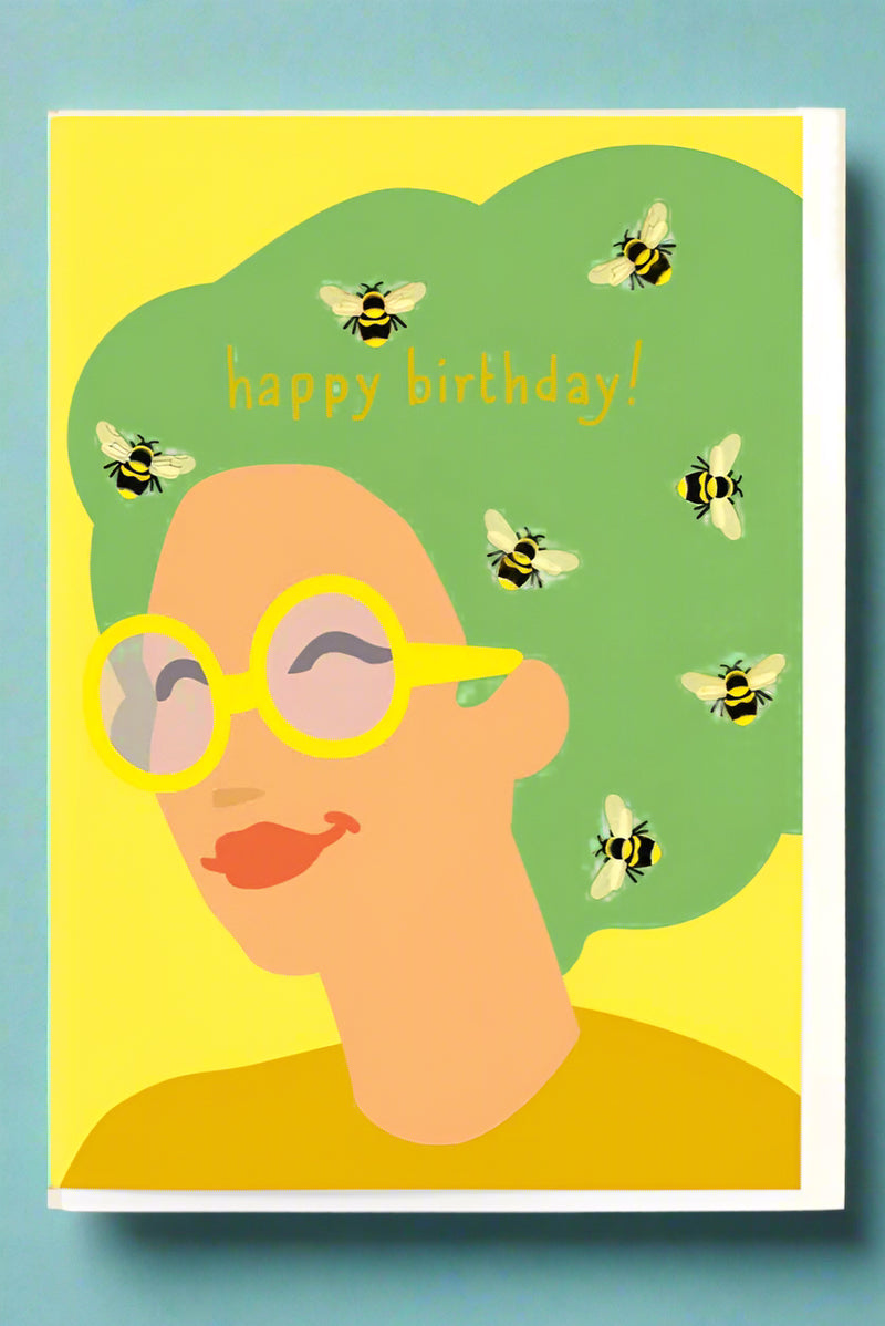 A greeting card with the image of a smiling woman with bees all in her hair and text that reads ‘happy birthday!’