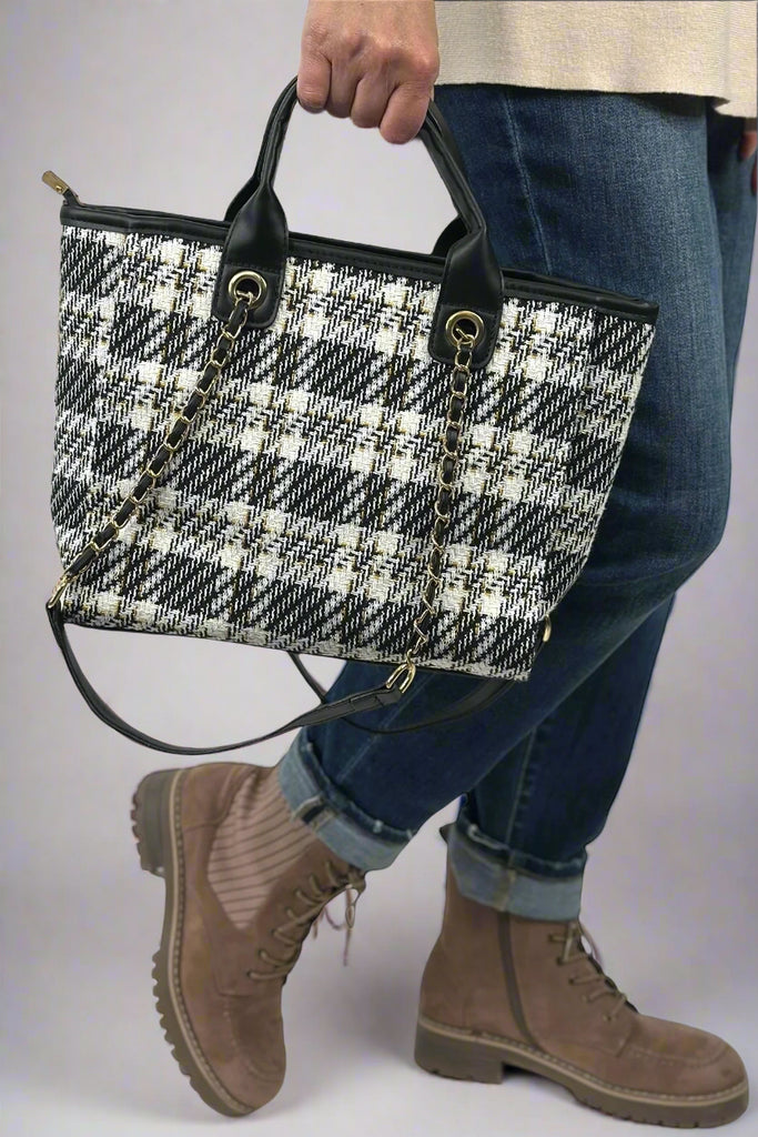 Model holds black and cream plaid tote bag with black, faux leather lining and gold chain detailing.