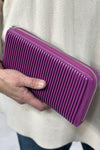 Model holds a bright, magenta/purple wallet constructed with vegan leather, featuring pleated detailing and a zipper closure.