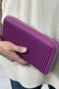 Model holds a bright, magenta/purple wallet constructed with vegan leather, featuring pleated detailing and a zipper closure.