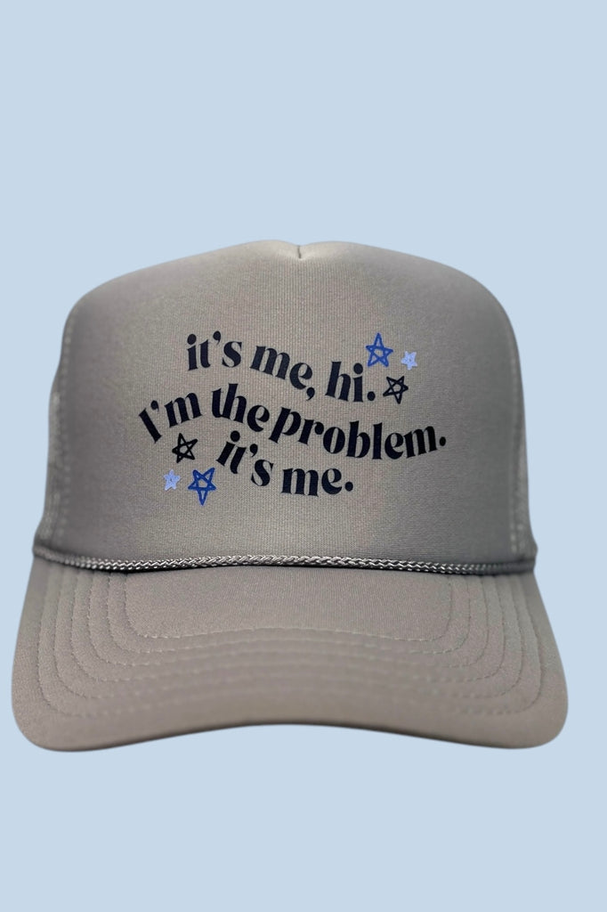 Grey foam front trucker hat with the phrase "It's me, hi. I'm the problem. It's me." and swirly black lettering. 