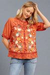 A women’s floral embroidered blouse in orange featuring a mandarin collar and half button front. Short sleeves, neckline, and hems with embroidered trim.