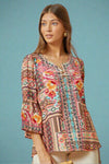 A women’s relaxed fit bell-sleeve blouse with split scoop neck featuring floral embroidery layered over a multicolored aztec patchwork print.